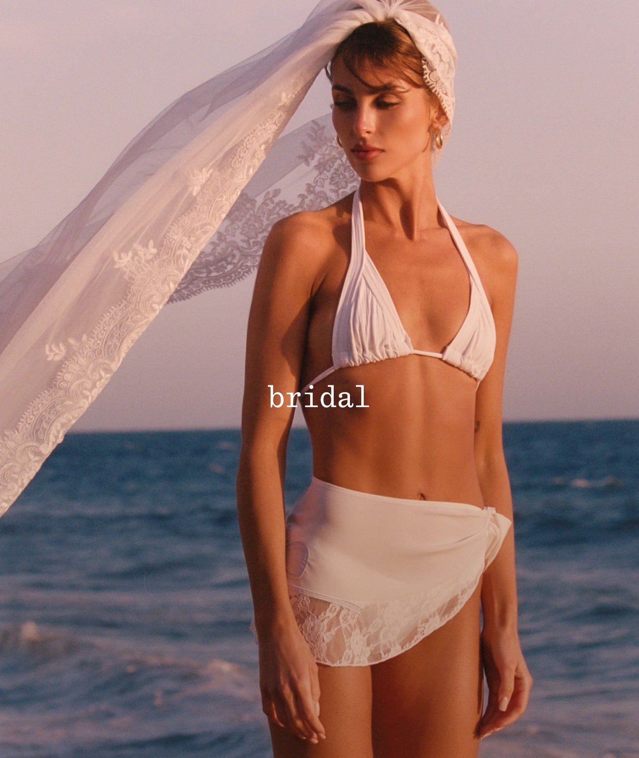 Paige standing on a beach wearing a white bridal two-piece swimsuit with a matching sheer lace headscarf. The background shows the blue ocean under a warm sunset sky.