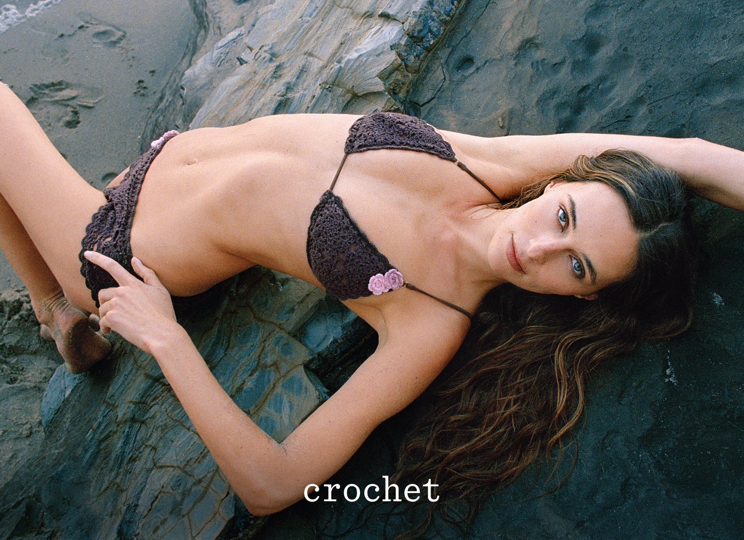 Devon reclines on dark rocks wearing a dark crochet bikini with small pink flower accents, with the word 'crochet' overlaid at the bottom.