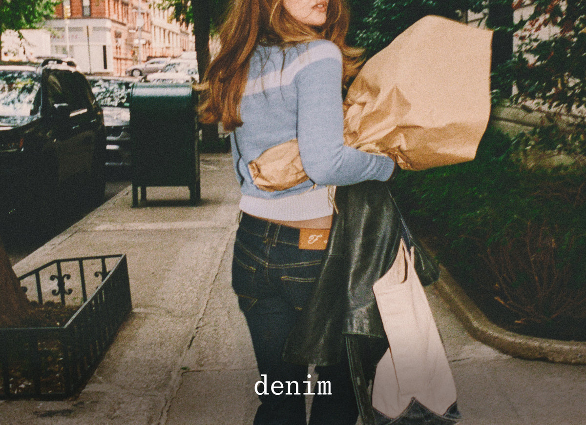A woman stands on a city sidewalk holding a large brown paper bag and a black leather jacket, wearing a light blue and gray striped long-sleeve top, dark blue jeans, and a brown leather belt with a visible 'denim' brand patch. The image has a warm, vintage filter and the word 'denim' is printed at the bottom.