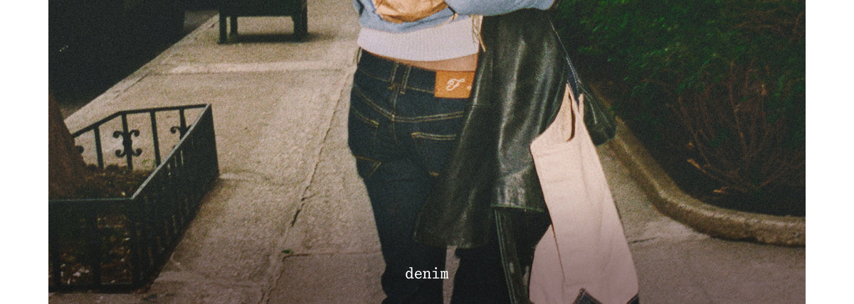 A person standing outdoors on a sidewalk wearing dark blue denim jeans, a light blue top, and carrying a black leather jacket and a light pink tote bag.