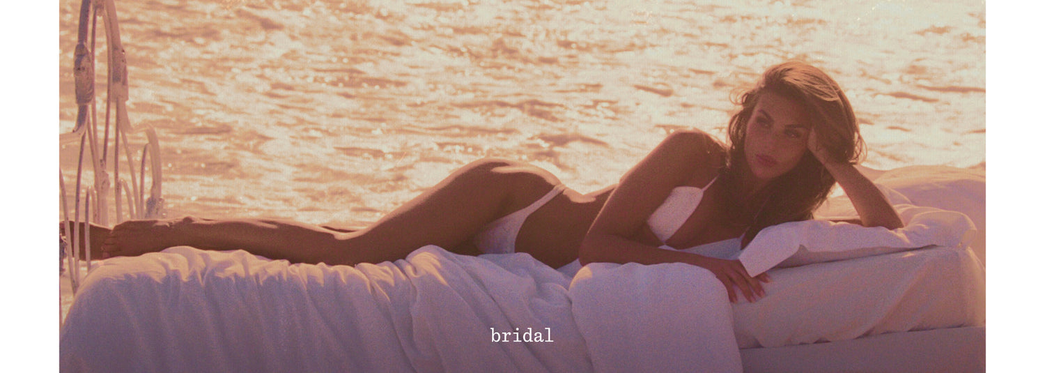 A woman reclining on a white bed or deck in warm golden-hour light, wearing a white bikini. The background shows shimmering water, and the word 'bridal' appears near the bottom center.