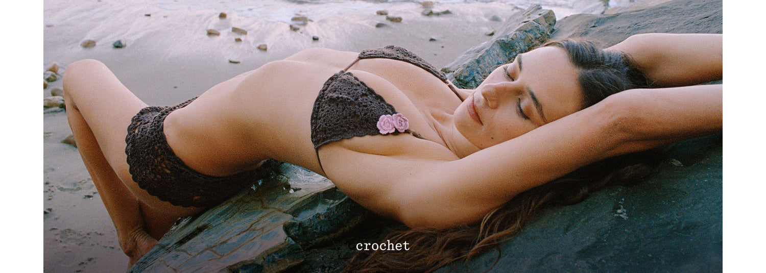 Devon reclining on a rock at the beach wearing a dark crochet bikini with a small pink flower accent, with the word 'crochet.' visible near the bottom.