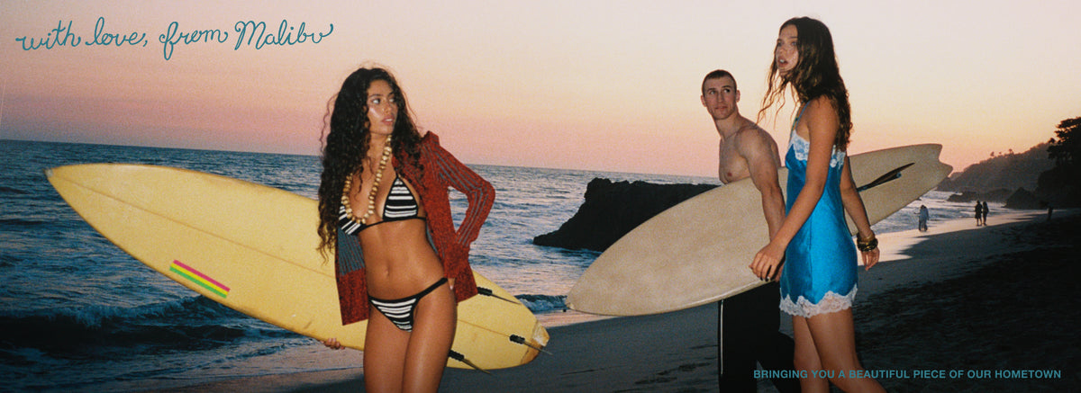 Three surfers standing on a beach at sunset, each holding a surfboard. The scene has a warm peach and orange sky, ocean waves, and rocks in the background.