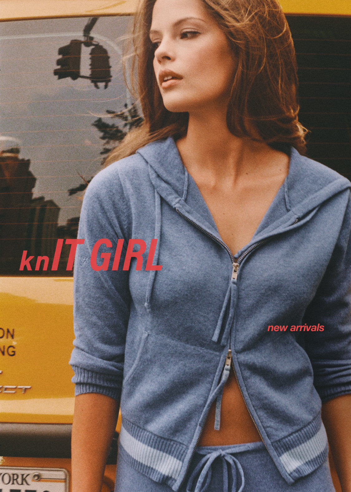 A woman wearing a blue zip-up hooded sweatshirt and matching drawstring bottoms, photographed in an urban setting with a yellow vehicle in the background. Red text overlays read 'knIT GIRL' and 'new arrivals'.