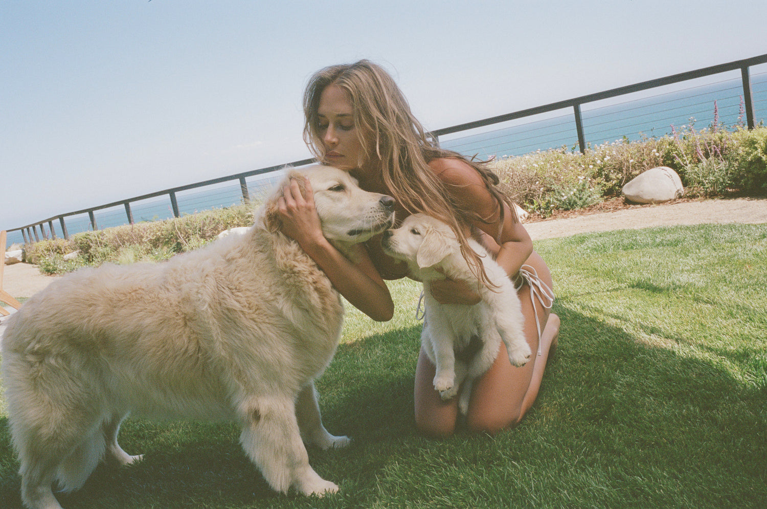 Francesca kneels on green grass outdoors by the ocean, next to a large cream-colored dog while holding a small white dog. A railing and blue sea are visible in the background.