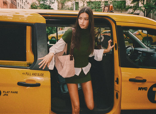 A woman stands in the open sliding door of a yellow NYC cab, wearing a dark green et with white ruffled sleeves and carrying a large blush pink tote bag. by Frankies Bikinis 