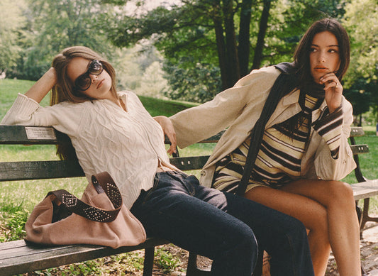 Two women sitting on a park bench, showcasing fashion apparel. One wears a cream cable-knit sweater and dark jeans with a beige handbag beside her, while the other wears a beige striped top and jacket. by Frankies Bikinis 
